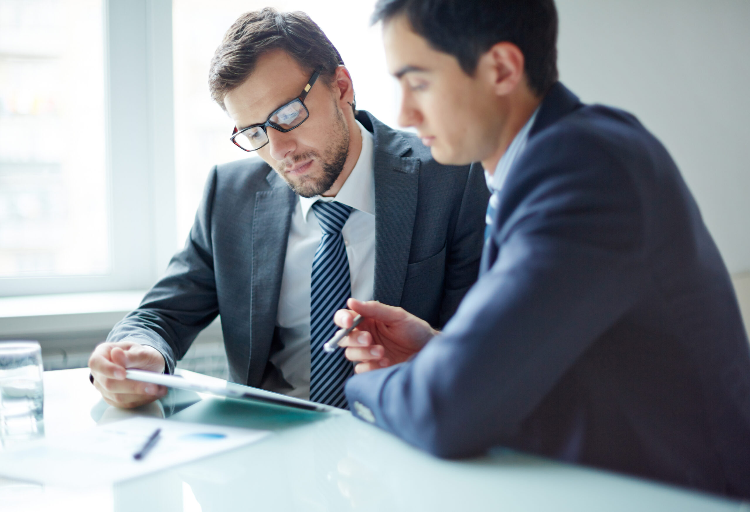 Two men in gray suits lean on a desk reviewing paperwork