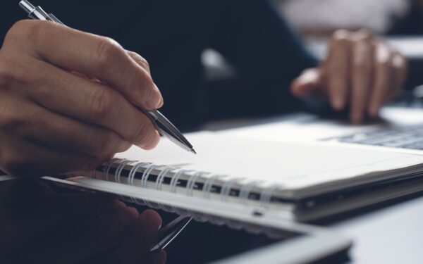 hand holding a pen over top of a notebook and ipad