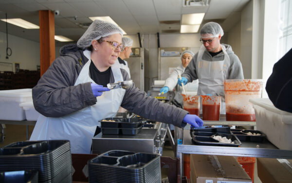 Woman and man in hairnets and aprons serving pasta into black pre-portioned containers