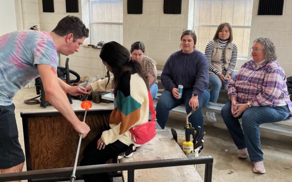 Honey Maung sits on bench holding a rod with an in progress glass blowing project on the end